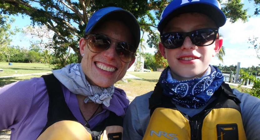 A parent and child wearing life jackets, sunglasses and hats smile at the camera.